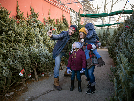 Mom and Dad with two young kids picking out christmas tree and taking silly selfie